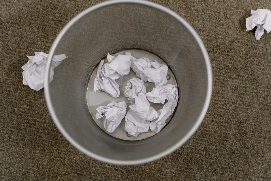 A cylindrical metal rubbish bin with a smooth, matte finish, positioned on a brown, textured carpeted floor. The bin contains several crumpled pieces of white paper, some partially submerged in a smal